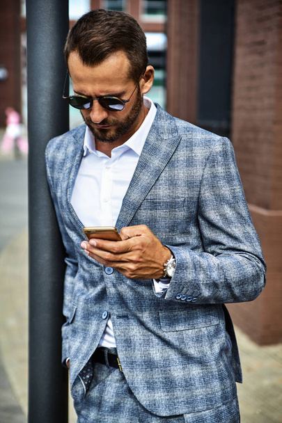 Portrait of sexy handsome fashion businessman model dressed in elegant checkered suit having business mobile conversation on smartphone on street. Metrosexual - Foto, immagini