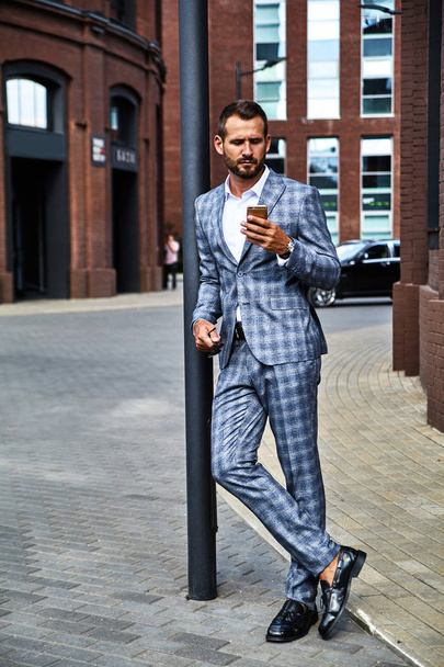 Portrait of sexy handsome fashion businessman model dressed in elegant checkered suit having business mobile conversation on smartphone on street. Metrosexual - Photo, Image