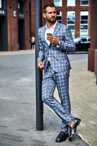 Portrait of sexy handsome fashion businessman model dressed in elegant checkered suit having business mobile conversation on smartphone on street. Metrosexual - Fotó, kép