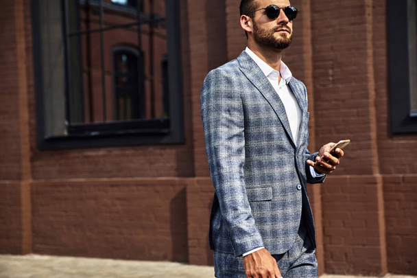 Portrait of sexy handsome fashion businessman model dressed in elegant checkered suit having business mobile conversation on smartphone on street. Metrosexual walking - Zdjęcie, obraz
