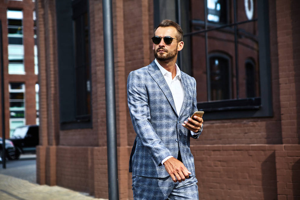 Portrait of sexy handsome fashion businessman model dressed in elegant checkered suit having business mobile conversation on smartphone on street. Metrosexual walking - 写真・画像