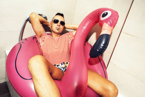 un joven caucásico con gafas de sol, un traje de baño y un anillo de natación en forma de flamenco rosa fingiendo que se está relajando en la bañera de su baño
 - Foto, Imagen