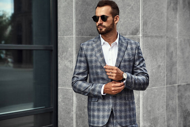 Portrait of sexy handsome fashion businessman model dressed in elegant checkered suit posing near gray wall on street background. Metrosexual - Fotó, kép