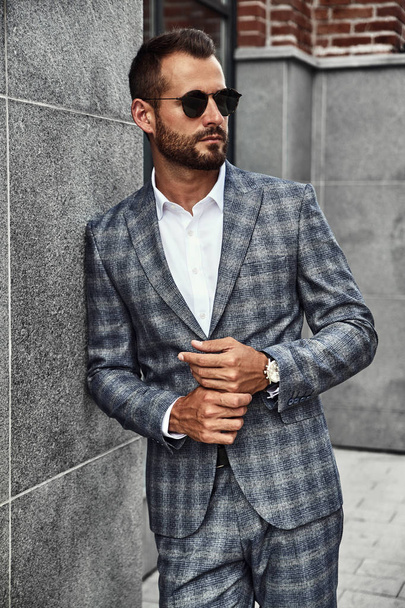 Portrait of sexy handsome fashion businessman model dressed in elegant checkered suit posing near gray wall on street background. Metrosexual - Foto, imagen