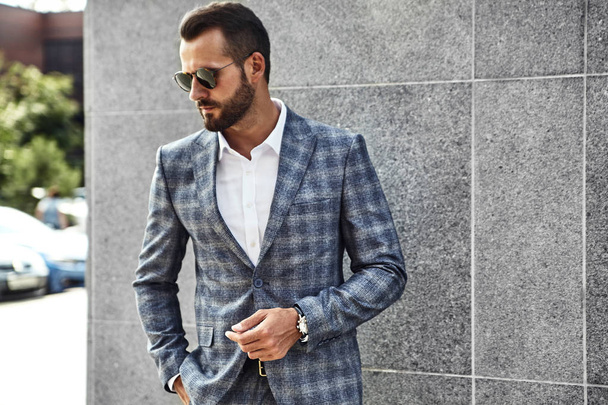 Portrait of sexy handsome fashion businessman model dressed in elegant checkered suit posing near gray wall on street background. Metrosexual - Φωτογραφία, εικόνα