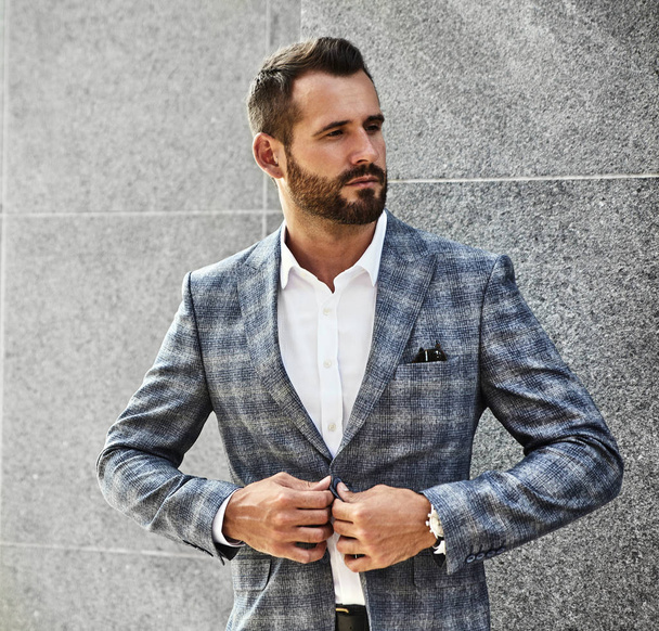 Portrait of sexy handsome fashion businessman model dressed in elegant checkered suit posing near gray wall on street background. Metrosexual - Foto, Bild