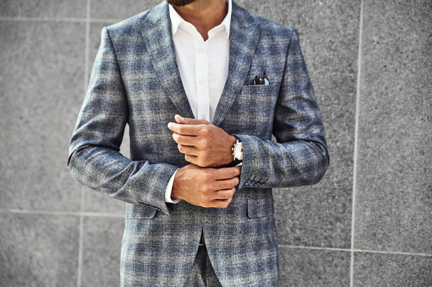 Fashion businessman model dressed in elegant checkered suit posing near gray wall on street background. Metrosexual with luxury watch on wrist - Foto, imagen