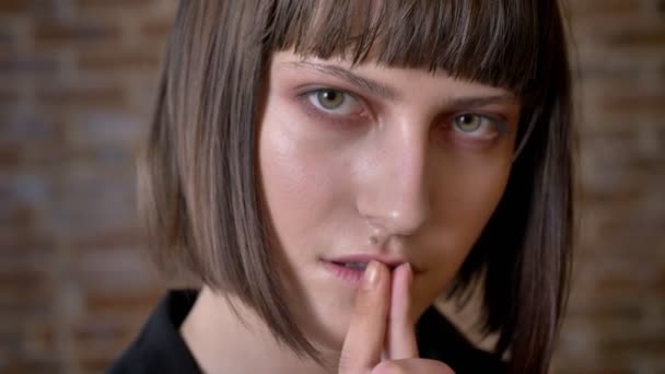 Young thoughtful woman with green eyes touching her lips looking at camera and sideways, brick background - Imágenes, Vídeo