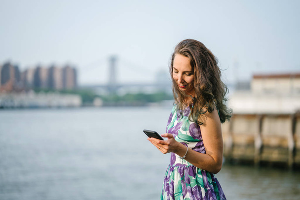 outdoor portrait of brunette woman with curly hair walking and holding smartphone on riverside on bridge background - Photo, Image