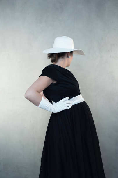 Schwangere im Retro-Look der 1950er Jahre mit weißem Hut und schwarzem Kleid. Wegschauen von der Kamera. - Foto, Bild