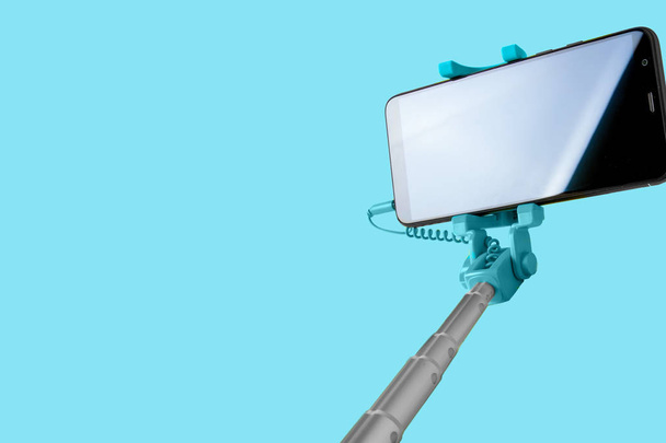 Monópodo para selfie con teléfono inteligente. Selfie stick con smartphone aislado sobre fondo azul
. - Foto, Imagen