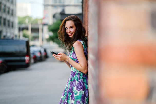beautiful brunette woman using smartphone standing at orange brick wall  - Фото, изображение