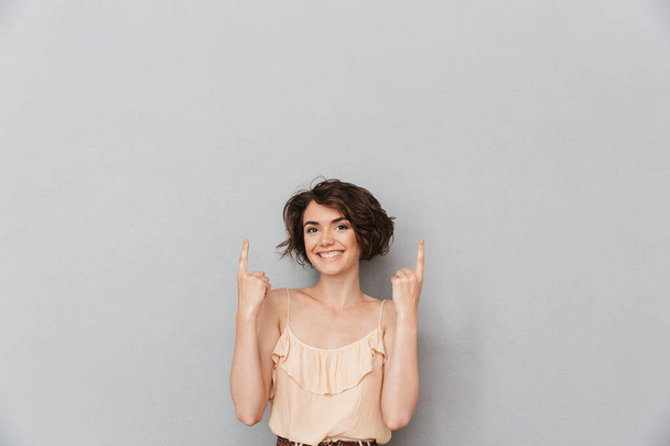 Portréja egy mosolygó fiatal nő, elszigetelt, felett szürke háttér copyspace ujjal mutatva - Fotó, kép