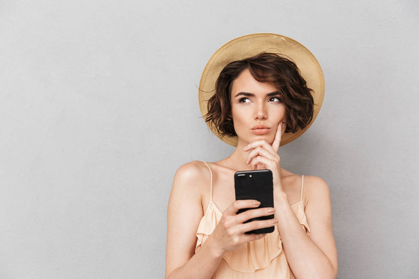 Close up portrait of a pensive young woman in summer hat holding mobile phone and looking away isolated over gray background - Фото, изображение