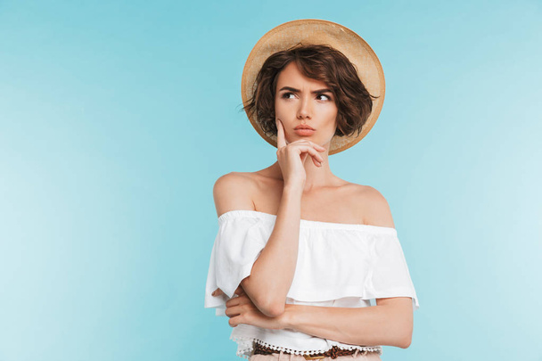 Portrait of a pensive young woman in summer hat looking away at copy space isolated over blue background - Photo, Image