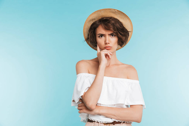 Portrait of a pensive young woman in summer hat looking at camera isolated over blue background - Photo, Image