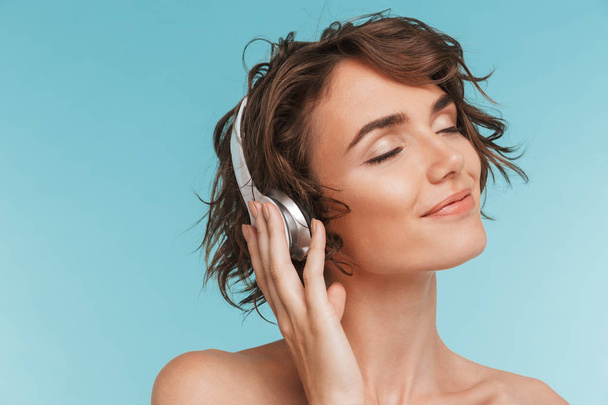 Close up portrait of a delighted young woman listening to music with headphones with eyes closed isolated over blue background - Φωτογραφία, εικόνα