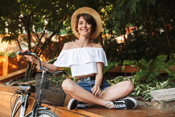 Iloinen nuori tyttö kesävaatteissa istuu puistossa polkupyörän kanssa
 - Valokuva, kuva