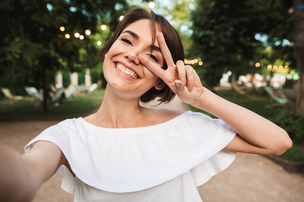 Smiling young girl showing peace gesture while taking a selfie at the park - Photo, image