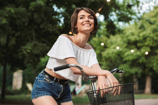 Smiling young girl in summer clothes sitting on a bicycle at the park - Photo, image