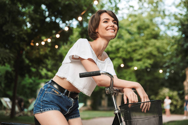 Pretty young girl in summer clothes sitting on a bicycle at the park - 写真・画像