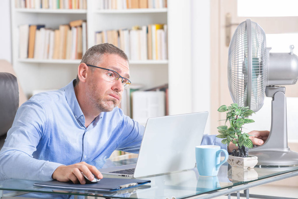 L'uomo soffre di calore mentre lavora in ufficio e cerca di rinfrescarsi dal ventilatore - Foto, immagini