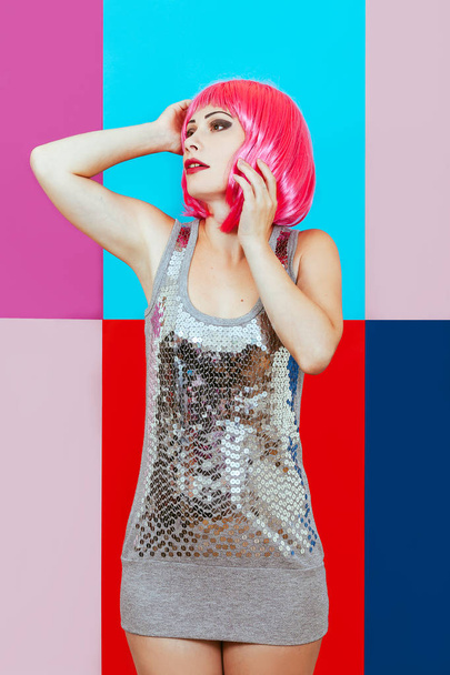 Beautiful girl with pink hair dressed in silver dress on a multicolored graphic background. A happy woman emotionally touches head her hands. - Photo, Image