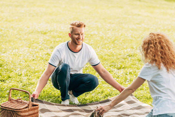 happy redhead couple holding plaid and looking at each other at picnic in park  - Photo, Image