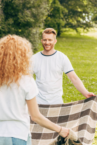 glückliches rothaariges Paar, das sich beim Picknick im Park anschaut und kariert hält  - Foto, Bild