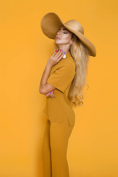 Красивая, сексуальная, элегантная женщина с светлыми волосами позирует в студии в элегантной желтой одежде и шляпе
. - Фото, изображение