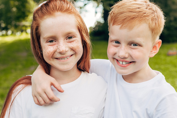 beautiful happy redhead siblings embracing and smiling at camera in park - Фото, зображення