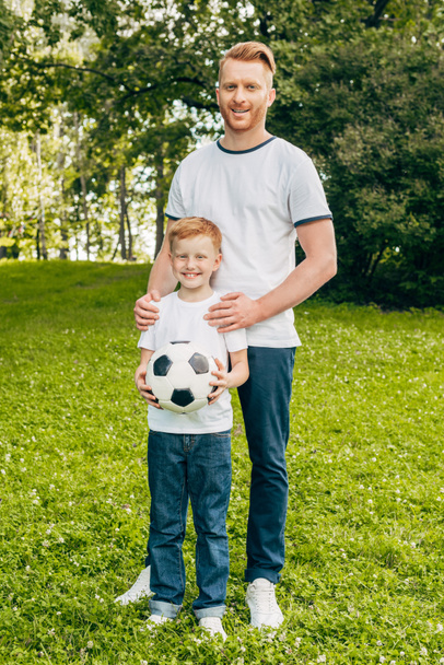 onnellinen isä ja poika jalkapallo seisoo yhdessä ja hymyilee kameralle puistossa
 - Valokuva, kuva