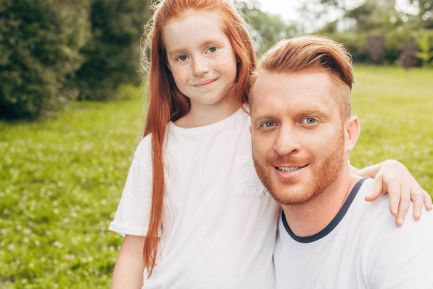 happy redhead father and daughter smiling at camera in park - Foto, immagini