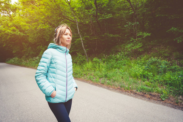 Portrait of an attractive sports girl blonde in a light running down jacket dressing bluetooth headphones with music or the sounds of nature while on a forest road - Photo, Image