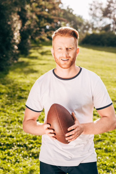 handsome man holding rugby ball and smiling at camera in park - Fotoğraf, Görsel