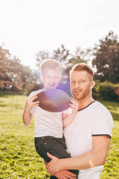 happy father and son holding rugby ball and smiling at camera in park - Valokuva, kuva