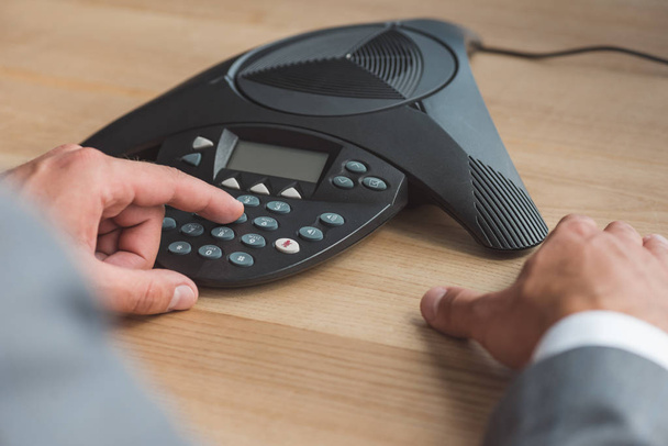 cropped shot of businessman pushing button of conference phone on wooden table at office - Photo, Image