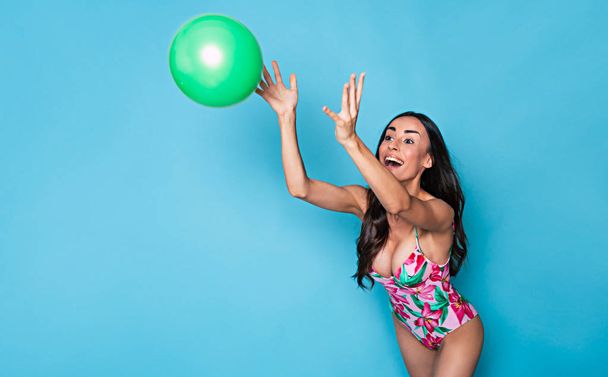 Smiling brunette woman in colorful swimsuit posing with green ball on blue background - Fotó, kép