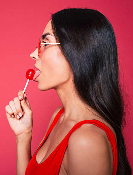 Young brunette woman with lollipop posing on red background - Foto, Imagem