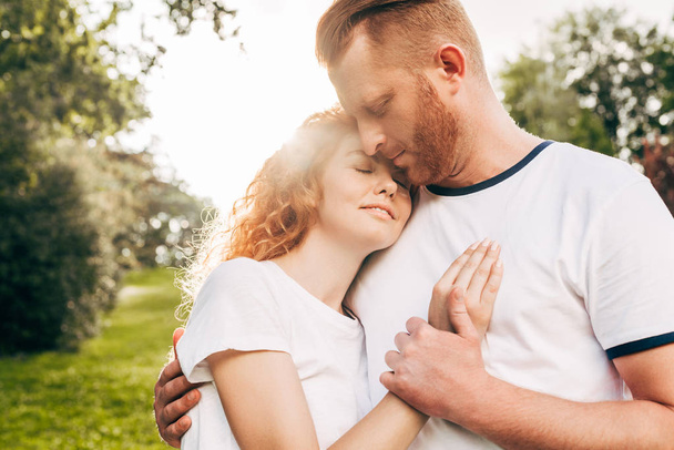 公園で一緒に立ったまま抱きしめる赤毛の幸せカップル - 写真・画像