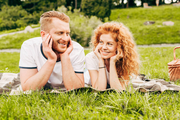 glückliches rothaariges Paar lächelt einander an, während es zusammen auf Plaid beim Picknick liegt - Foto, Bild