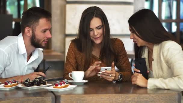 Cheerful teen hipster guy looking at camera while showing her girlfriend video on smartphone, romantic couple in love spending time together on cafe terrace viewing funny photos on mobile phone - Footage, Video