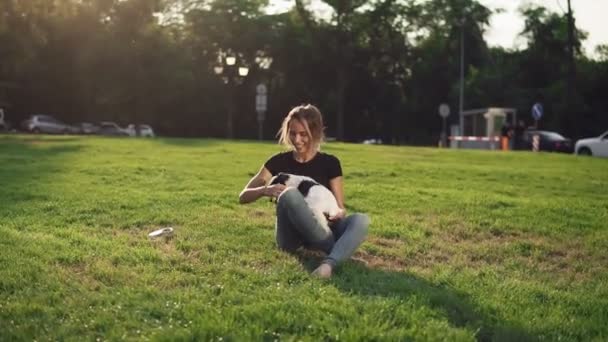 Beautiful young woman in casual sitting on grass with her dog pet and smiling. Owner with lovely little dog outdoors. Blue sky and sun in background and green grass - Video, Çekim