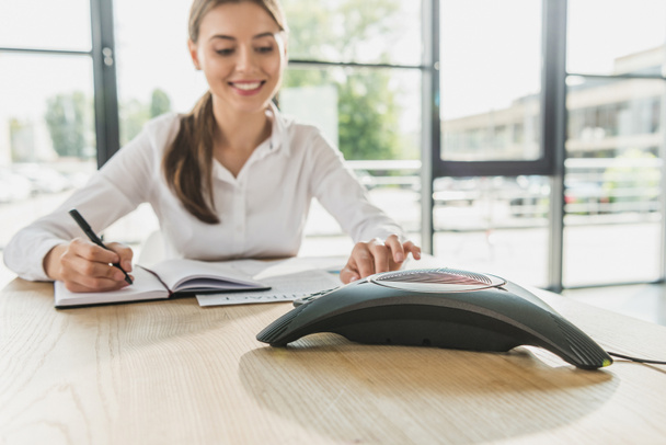 молодая счастливая деловая женщина делает бумажную работу и нажимает кнопку конференц-телефона на столе в офисе
 - Фото, изображение
