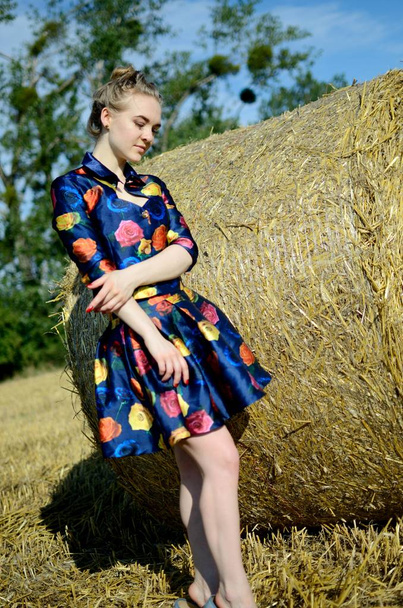 Modelka, venkovní focení, zemědělské půdy v létě. Dívka v barevných šatech stojí před roll sena. - Fotografie, Obrázek