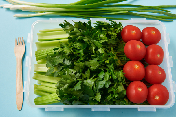 Close-up van vork en voedsel container vol verse tomaten, peterselie en selderij op blauwe achtergrond - Foto, afbeelding
