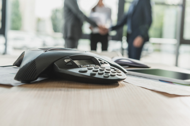 close-up shot of conference phone with blurred business people shaking hands on background at modern office - Fotografie, Obrázek