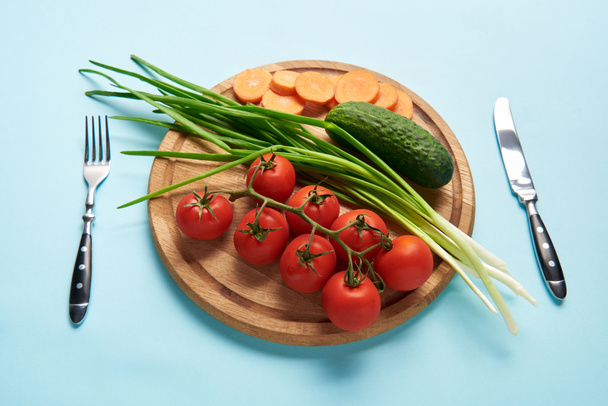 close up view of cutlery and assorted fresh vegetables on wooden cutting board on blue backdrop - Foto, imagen