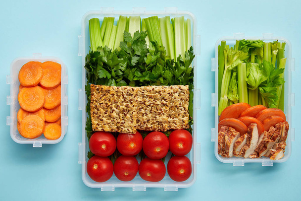 top view of arrangement of food containers with fresh healthy vegetables, cookies and meat isolated on blue - 写真・画像