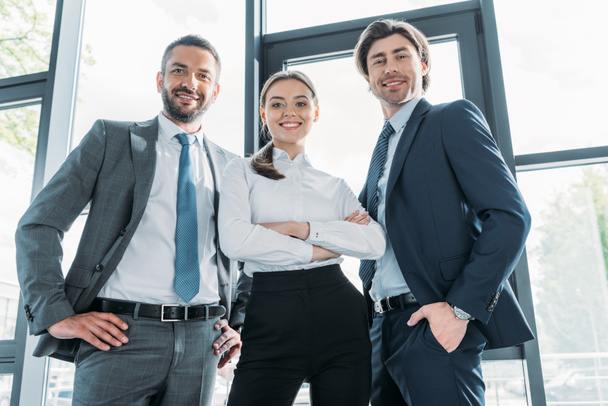 вид снизу группы улыбающихся молодых бизнесменов, стоящих в современном офисе и смотрящих в камеру
 - Фото, изображение